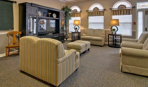 A cozy living room with multiple upholstered chairs and sofas arranged around a large black entertainment center with a flat-screen TV. The room has three arched windows with blinds and valances, two side tables with lamps, and a carpeted floor.