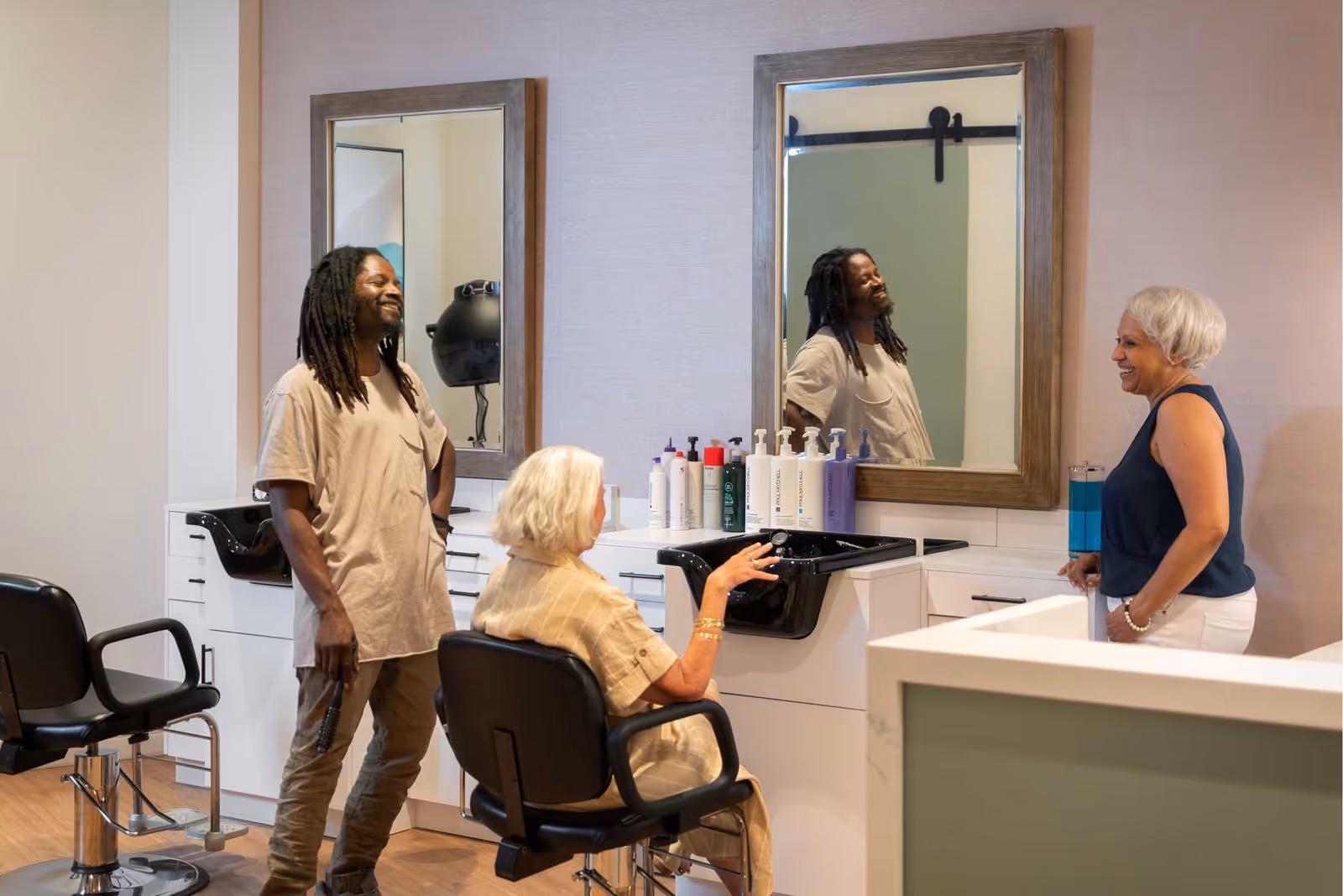 A hair salon area with two large mirrors mounted on the wall above black sinks and white cabinetry. A man with dreadlocks stands smiling next to a seated elderly woman with white hair, while another woman with short gray hair stands nearby, all engaged in conversation.