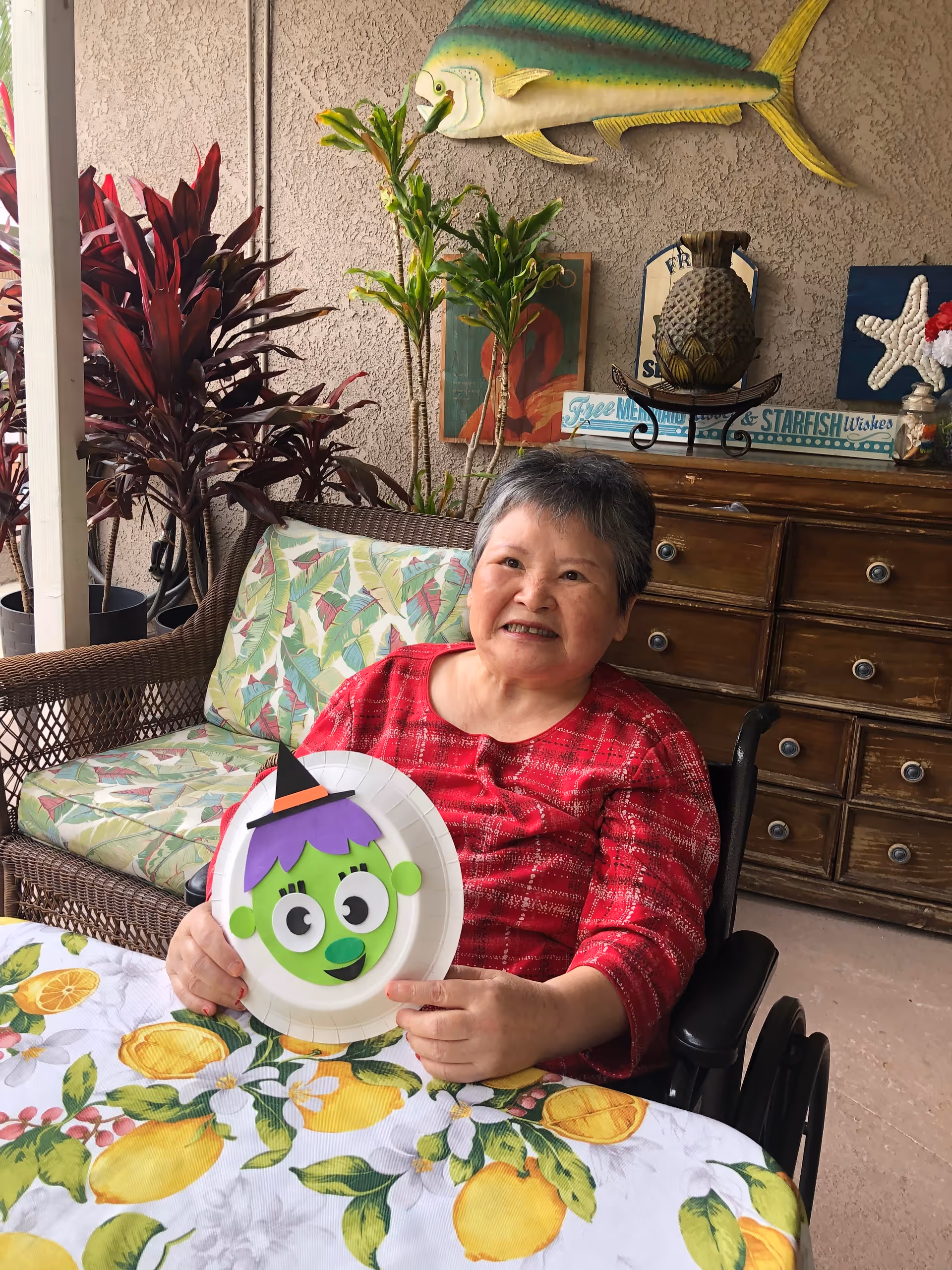 A person in a wheelchair sits at a table on a decorated patio holding a paper plate craft of a green cartoon face.