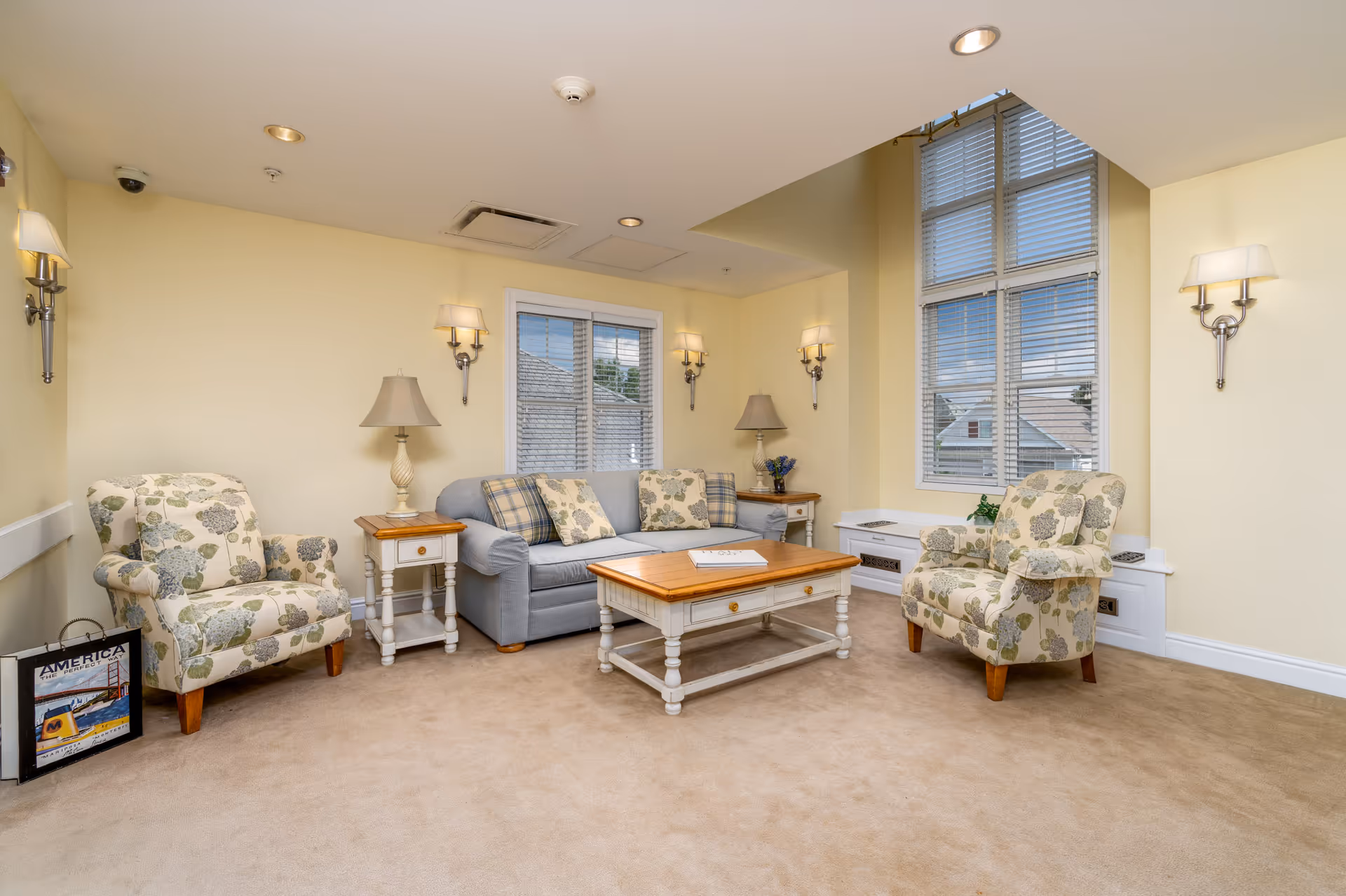 A cozy living room with beige walls and carpet, featuring a light blue sofa with patterned cushions, two floral armchairs, a wooden coffee table, two side tables with lamps, wall-mounted sconces, and large windows with blinds letting in natural light.