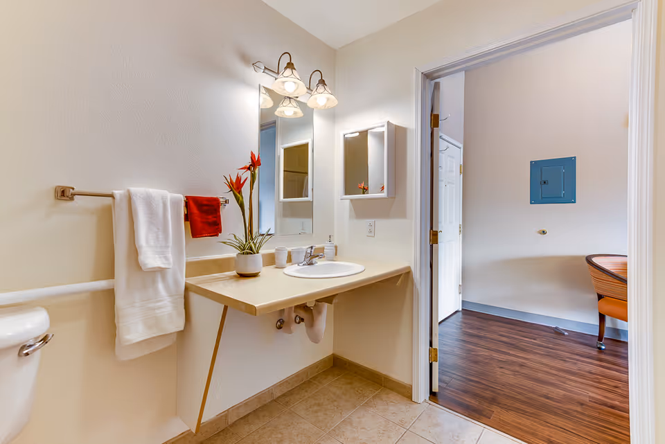 A clean bathroom with beige tiled floor and cream-colored walls. There is a countertop with a sink, a mirror above it, and a wall-mounted cabinet. Two towels, one white and one red, hang on a towel rack next to the sink. A potted plant with red flowers is placed on the countertop. The bathroom opens into another room with wooden flooring and a chair partially visible.