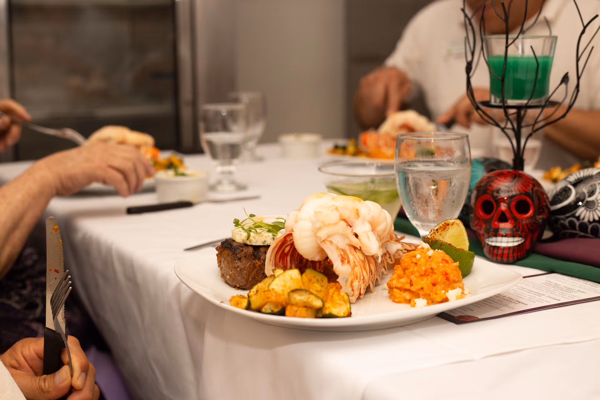 A close-up of a plated meal featuring lobster tail, a piece of steak topped with herb butter, roasted zucchini, and a serving of seasoned rice with a lime wedge on the side. The plate is on a white tablecloth with a glass of water and decorative skull candle holders nearby. People are seated at the table in the background, eating.