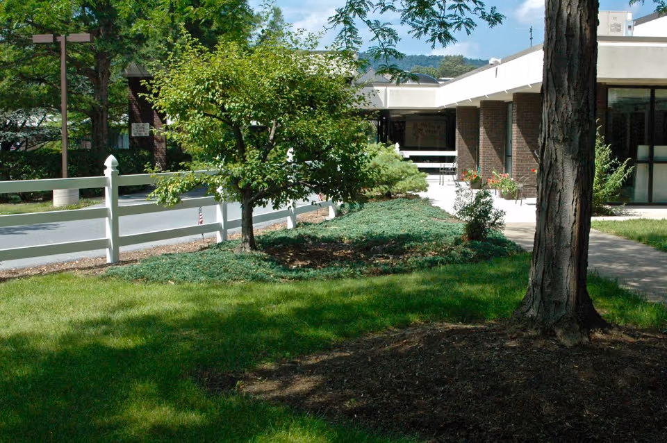 Outdoor view of a senior living community showing a green lawn with trees and shrubs, a white fence along a road, and a building with large windows and a covered walkway in the background under a partly cloudy sky.