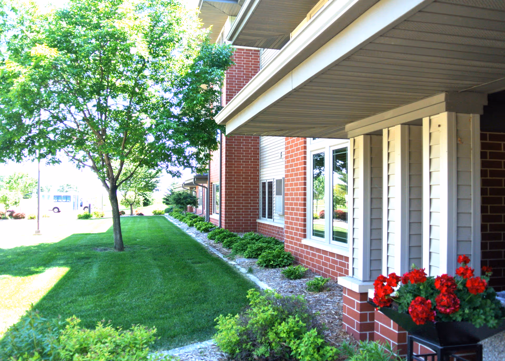 Exterior view of a brick-and-siding senior living building with a covered walkway, green lawn, a tree, and red flowers.