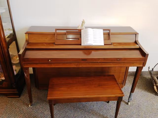 A wooden upright piano with a matching wooden bench in front of it. There is an open sheet music book on the piano's music stand. The piano is placed on a carpeted floor next to a wooden cabinet and a basket with some items inside.