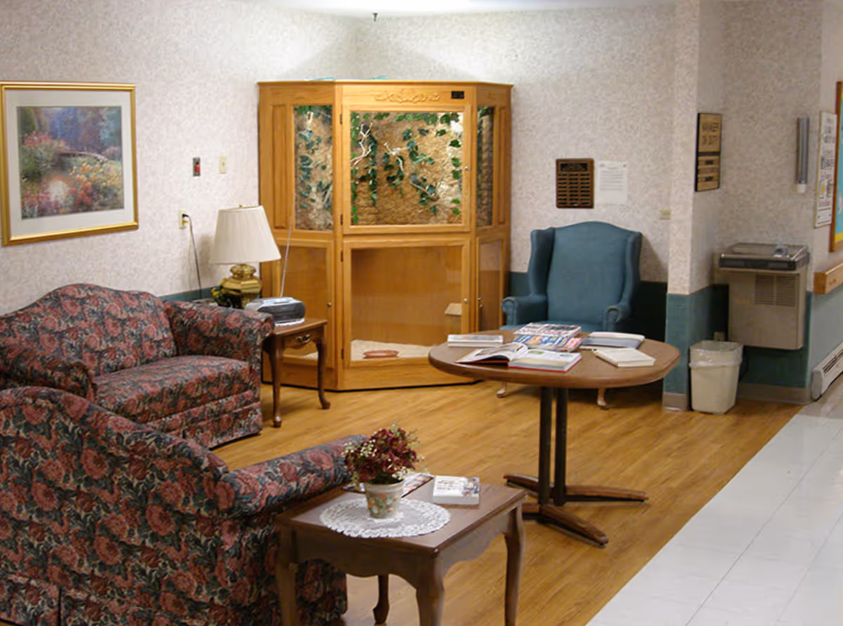 A senior living seating area with floral sofas, an armchair, a round table with magazines, side tables and a wooden display cabinet.