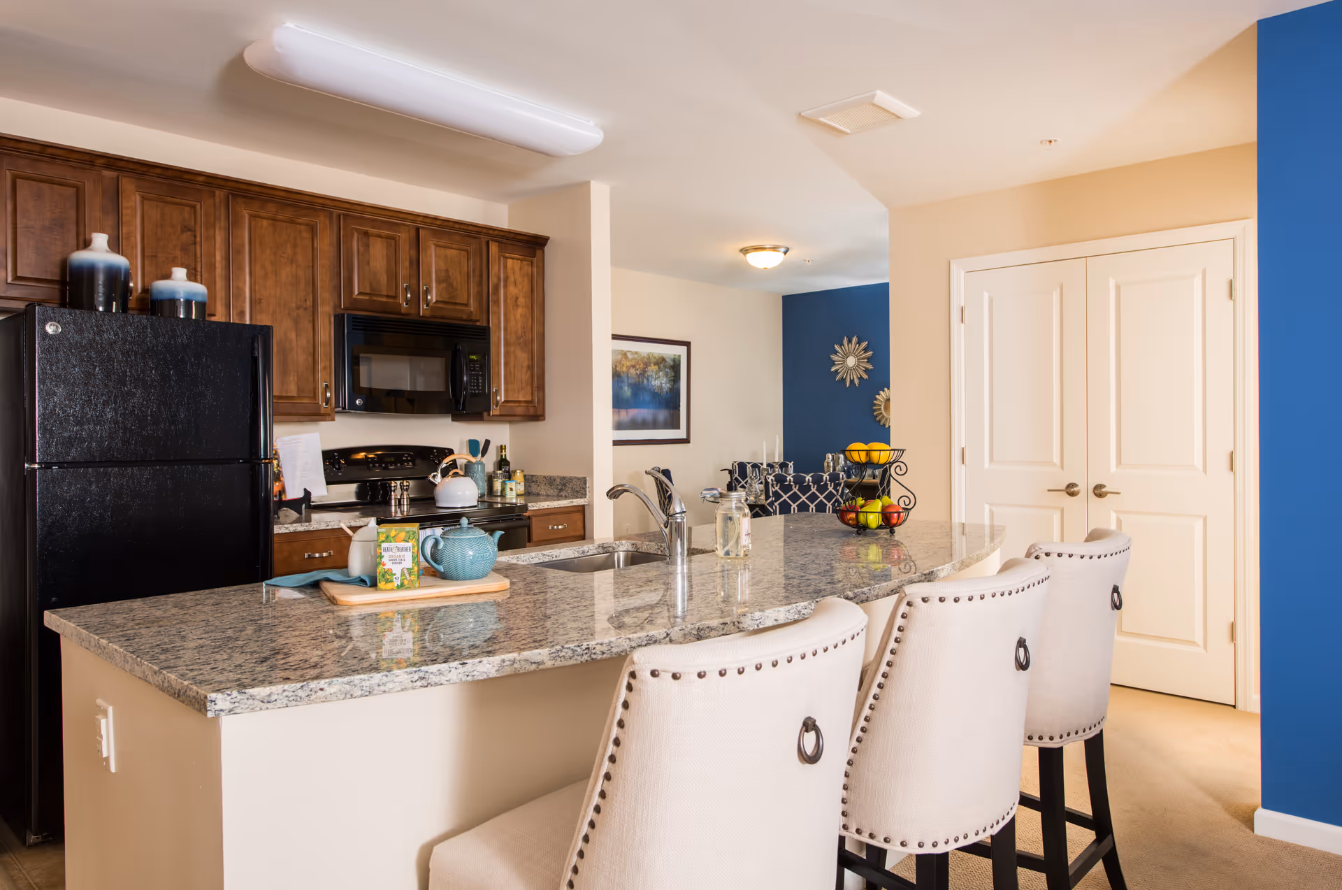 Open kitchen with a granite island, three upholstered bar stools, dark appliances and wooden cabinets in a senior living apartment.