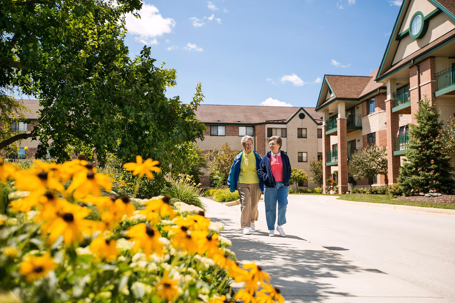 Two elderly women walking and talking on a sidewalk next to a senior living community building with yellow flowers and green trees in the foreground under a blue sky with some clouds.
