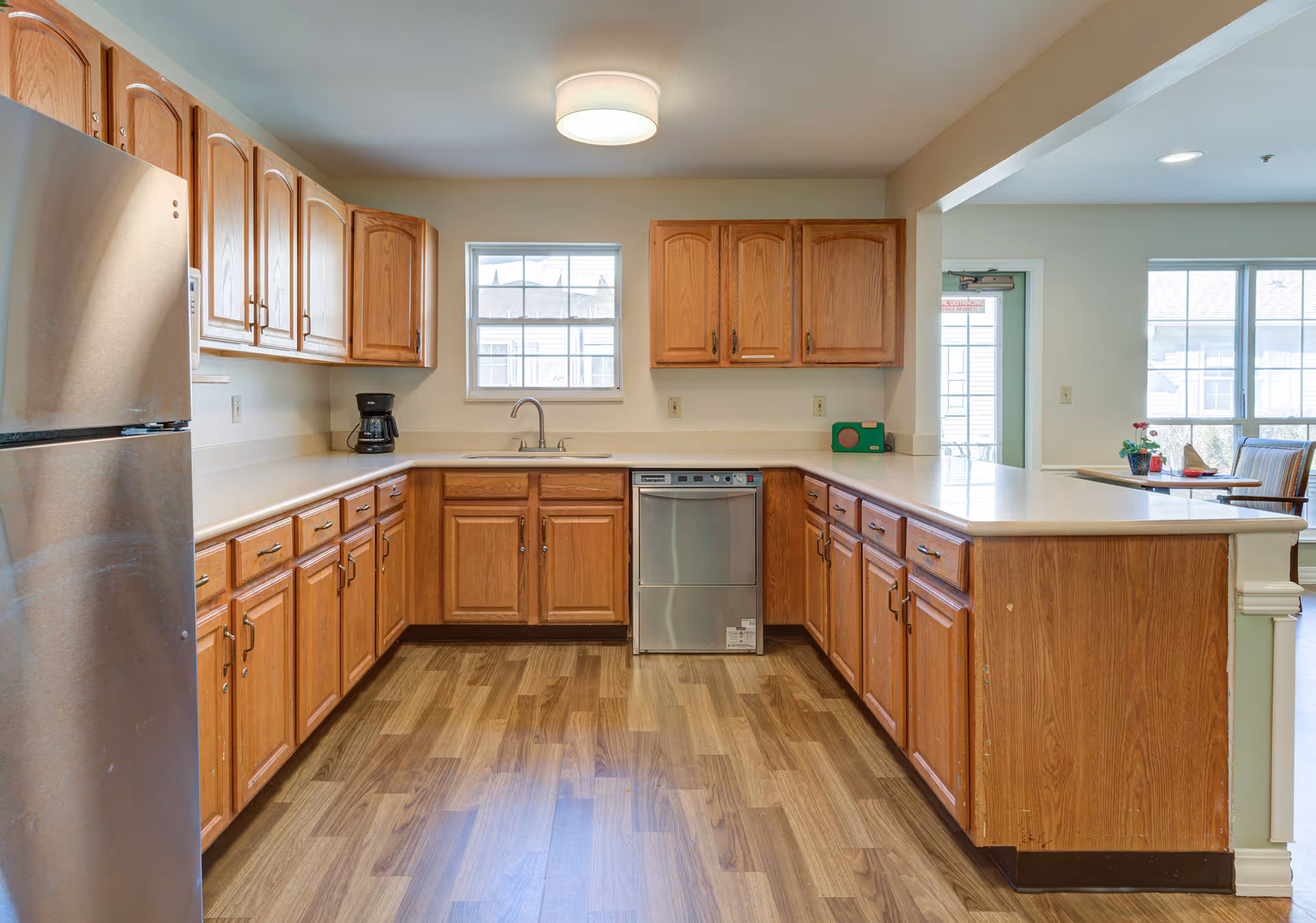 A spacious kitchen with wooden cabinets and drawers, a stainless steel refrigerator on the left, a dishwasher under the counter, a coffee maker on the counter, and a window above the sink letting in natural light. The kitchen has wood-patterned flooring and a light fixture on the ceiling. To the right, there is a partial view of a dining area with a table and chairs near windows.