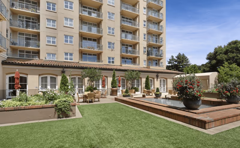 Outdoor courtyard area of a senior living facility with green lawn, brick planters, potted plants with red flowers, patio tables and chairs with umbrellas, and a multi-story building in the background under a clear blue sky.
