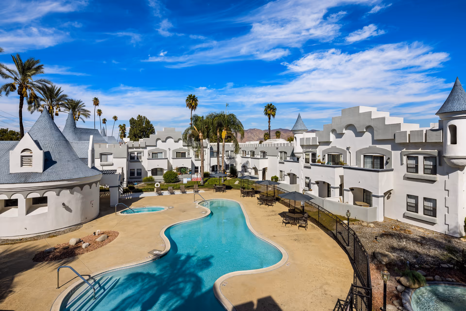 Outdoor view of a senior living community named Camelot featuring a uniquely designed white building with castle-like turrets surrounding a kidney-shaped swimming pool. The area includes lounge chairs, tables with umbrellas, palm trees, and a clear blue sky with scattered clouds.