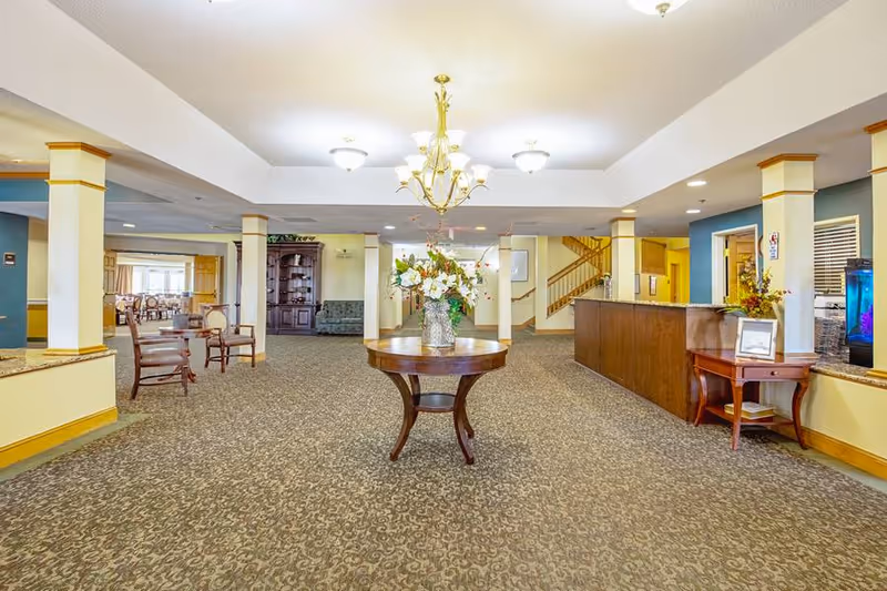 A spacious and well-lit interior common area of an assisted living facility featuring a round wooden table with a floral arrangement in the center, carpeted floor, multiple columns, a wooden reception desk on the right, chairs and tables in the background, and a staircase leading upstairs.