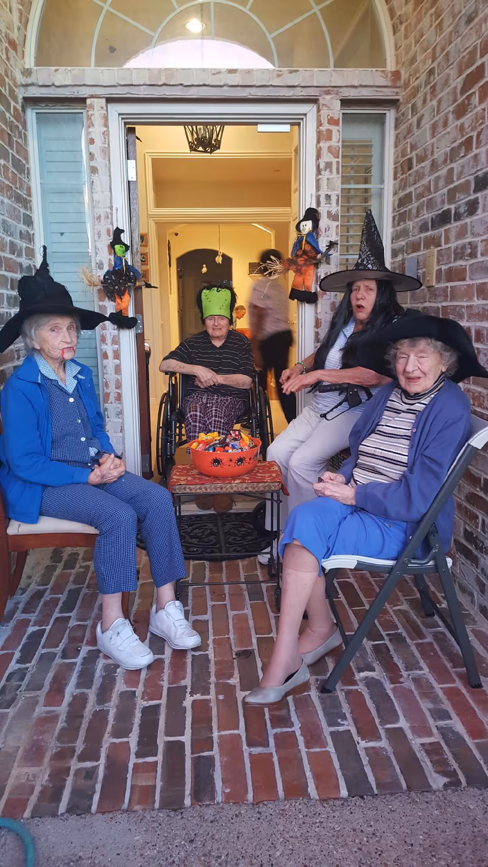 Four elderly women sitting outside on a brick patio in front of a doorway, dressed in Halloween costumes with witch hats and a Frankenstein hat. There is a small table with a bowl of candy in the center. The doorway behind them shows a hallway with a person walking inside.