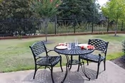 Metal patio table with two chairs and plates of snacks on a concrete patio overlooking a grassy yard and iron fence.