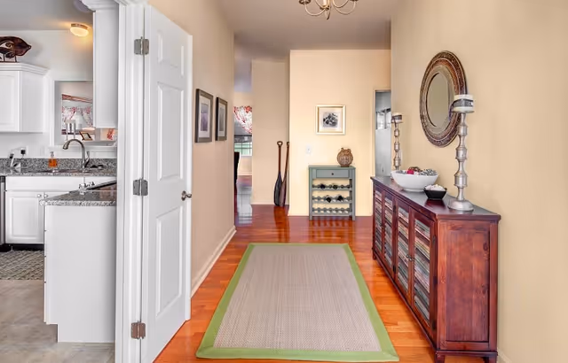 A hallway with light beige walls and wooden flooring featuring a beige rug with a green border. On the right side, there is a wooden cabinet with glass doors, decorated with a round mirror, two tall candle holders, and a bowl with decorative items. At the end of the hallway, there is a small wine rack and two oars leaning against the wall. To the left, an open door reveals a kitchen area with white cabinets and a granite countertop.