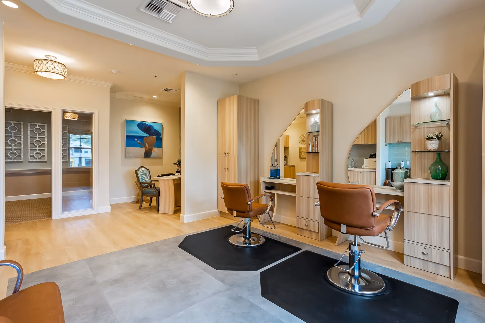Interior view of a salon area in a senior living facility with two brown salon chairs in front of large mirrors and wooden cabinetry. The room has light wood flooring, a small desk with a chair in the background, and soft lighting with a decorative ceiling fixture.
