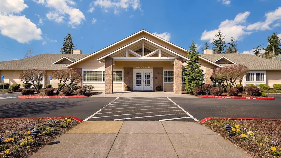 Front exterior view of a single-story senior living facility building with a peaked roof, beige siding, stone pillars at the entrance, and landscaped bushes and flowers along the walkway. The sky is blue with some clouds.