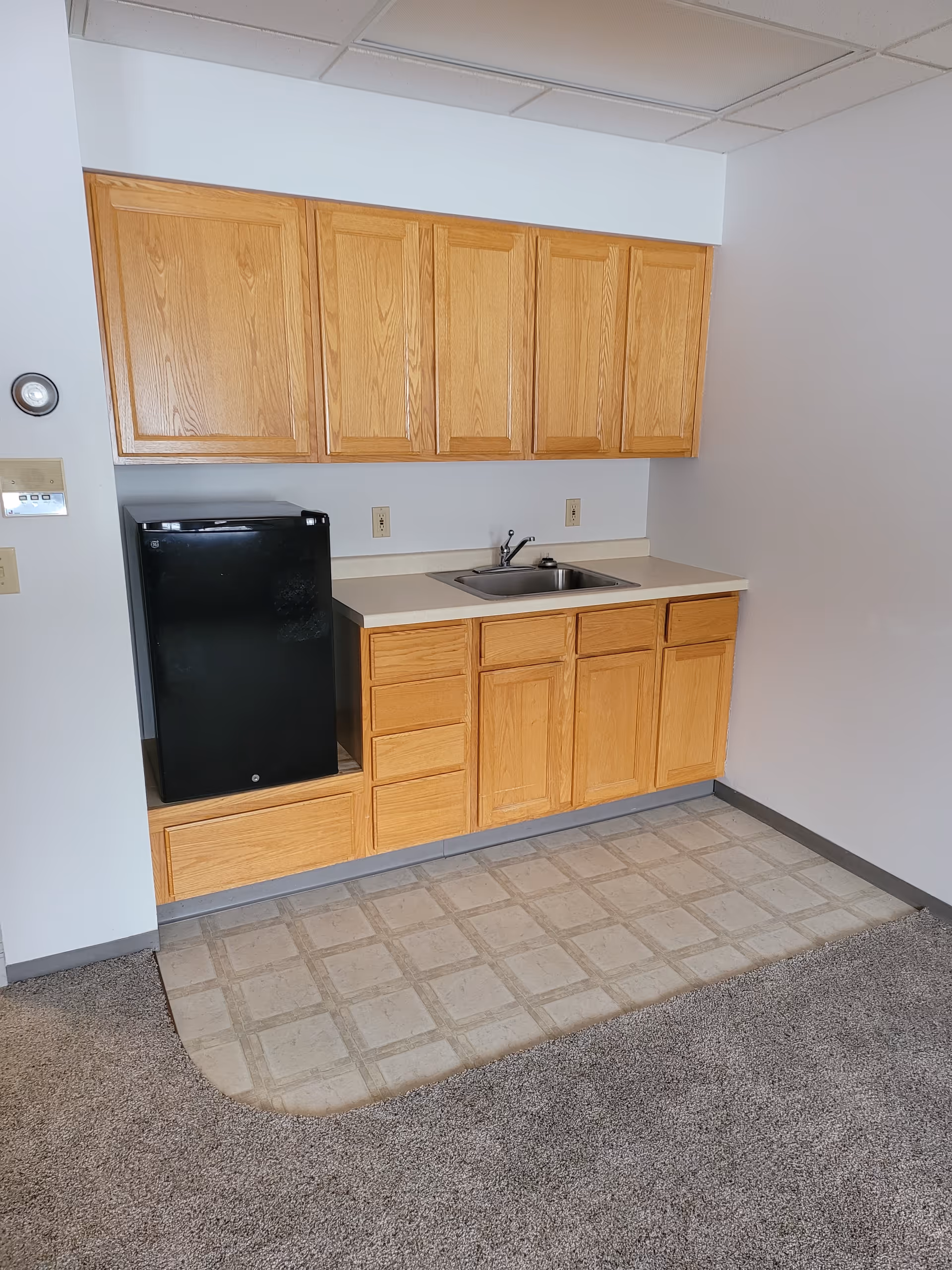 Small kitchenette with oak cabinets, a countertop sink, and a compact black refrigerator above vinyl flooring that meets carpet.