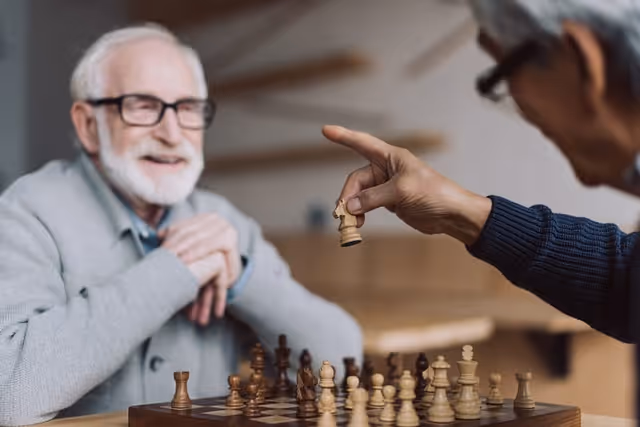 Two elderly men playing chess indoors, one man is holding a chess piece while the other watches with a smile.