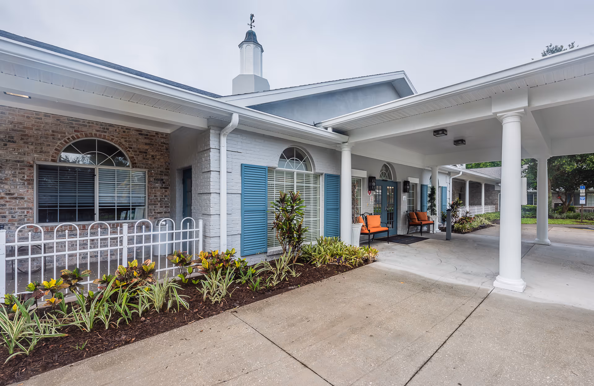 Exterior view of The Colonnade at Carrollwood senior living facility entrance with white columns, blue shutters, brick and painted walls, orange cushioned benches, and landscaped plants along the walkway.