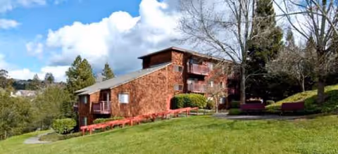 Exterior view of brick senior living building on a grassy hill with trees and red benches.