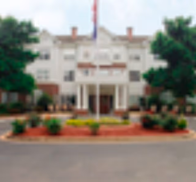 Front exterior view of a multi-story senior living facility building with a covered entrance, flagpole, and landscaped area with shrubs and flowers in front.