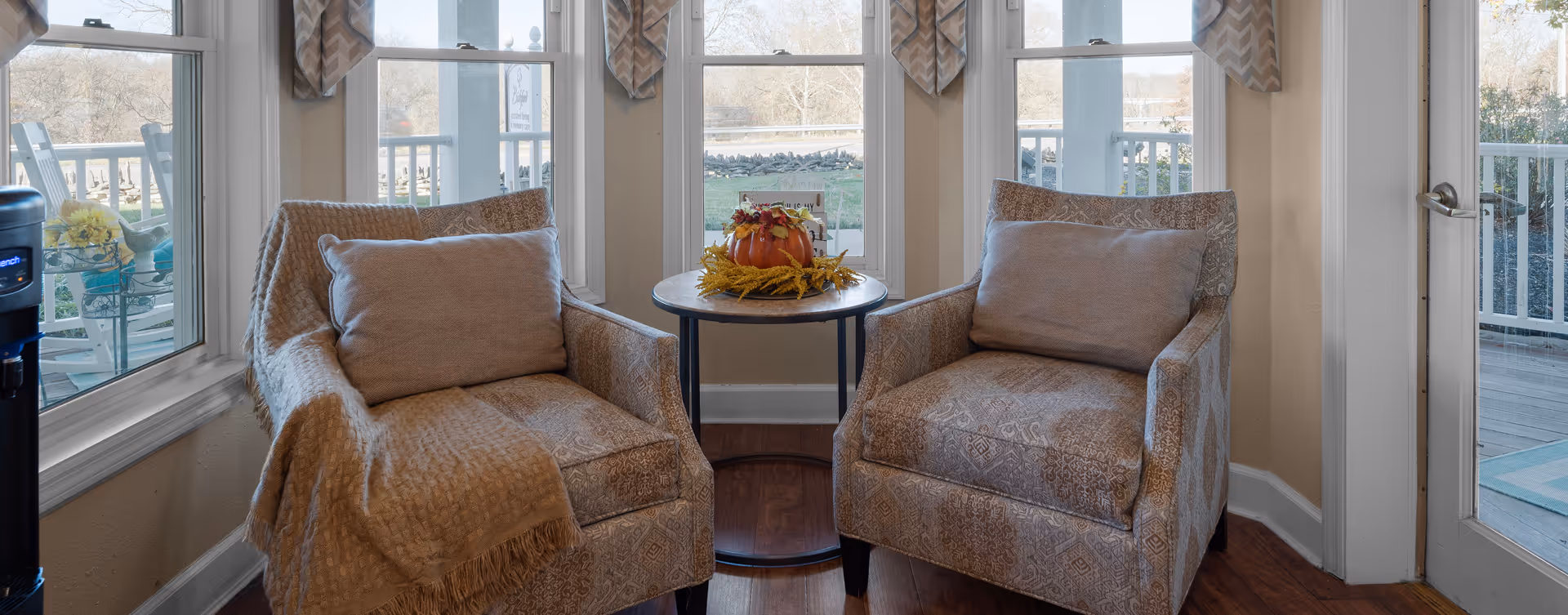 Two patterned armchairs with cushions are positioned around a small round table that has a fall-themed centerpiece. The seating area is next to large windows and a glass door that lead to an outdoor deck with a view of a grassy area and trees.