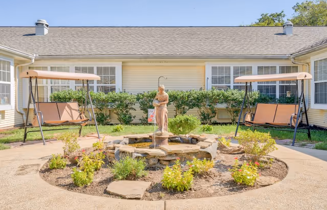 Outdoor courtyard area with a circular stone fountain featuring a statue in the center, surrounded by small plants and flowers. Two brown cushioned swing benches with canopies are positioned on either side of the fountain. The courtyard is enclosed by a yellow building with multiple windows and green shrubbery along the walls.
