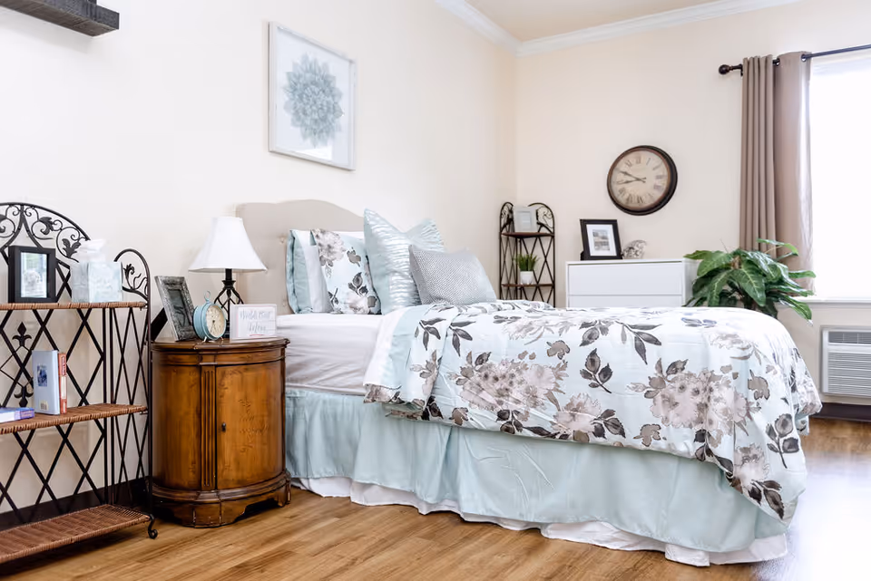 A bright and cozy bedroom with a bed dressed in floral and pastel bedding, multiple pillows, a wooden nightstand with a lamp, clock, and picture frames, a metal shelf with books and tissues, a white dresser with framed pictures and a clock on the wall above it, a large green plant near a window with beige curtains, and light wood flooring.