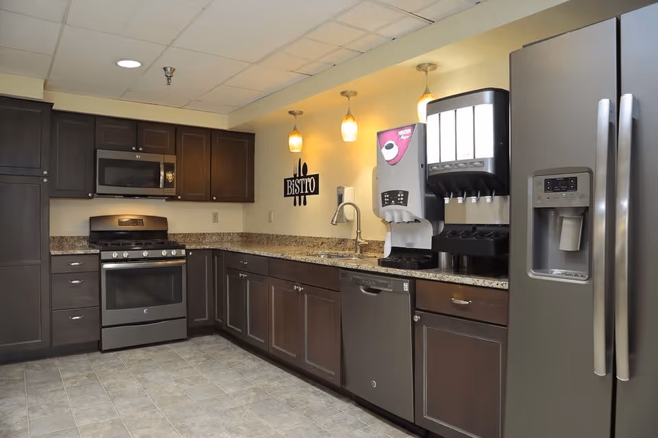 Modern kitchen area with dark wood cabinets, stainless steel appliances including a refrigerator, oven, microwave, and dishwasher. There is a granite countertop with a sink and two beverage dispensers on the right side. Three pendant lights hang above the countertop, and a wall decoration reads 'BISTRO'.