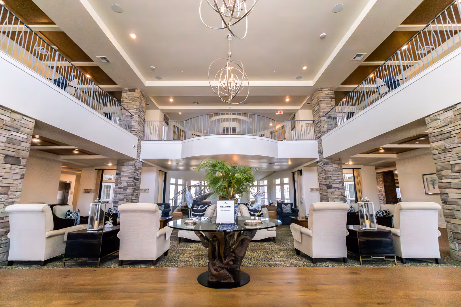 Spacious and elegant senior living facility common area with high ceilings, modern chandeliers, stone pillars, and a mezzanine balcony. The room is furnished with comfortable armchairs arranged around a central glass table with decorative items and a plant. Large windows allow natural light to fill the space.