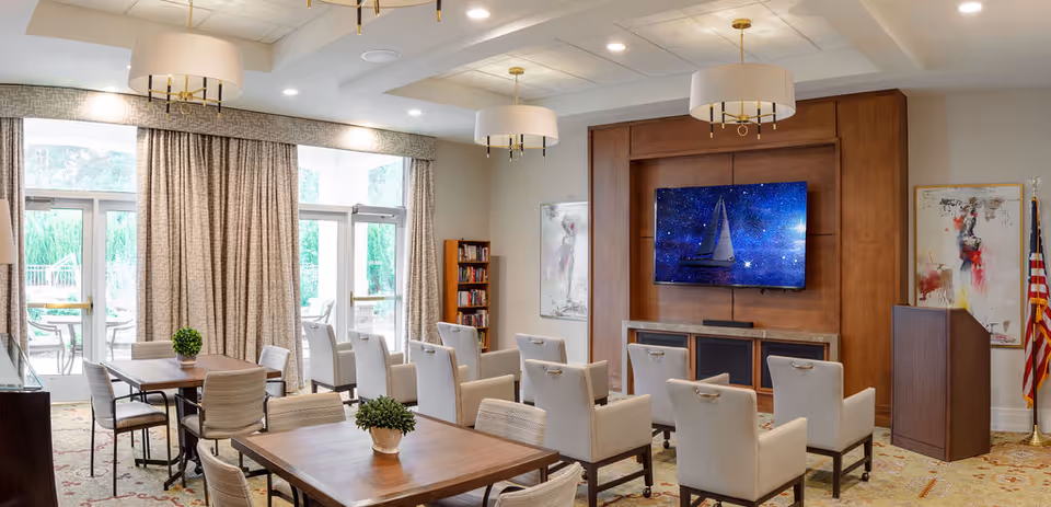 A well-lit common area with multiple tables and chairs arranged for seating. There are beige upholstered chairs facing a large flat-screen TV mounted on a wooden panel. The room features large windows with curtains, two modern ceiling light fixtures, a bookshelf, and abstract artwork on the walls. An American flag stands in the corner near a wooden podium.