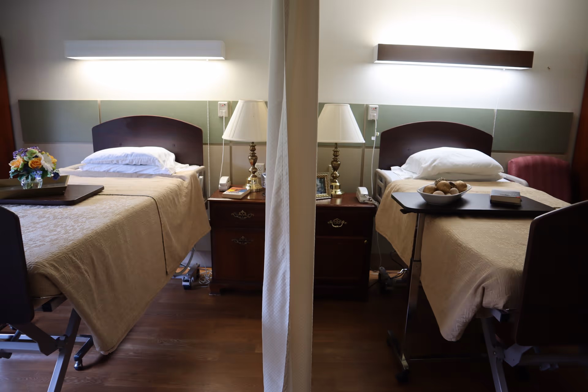 A shared room in a senior living facility with two hospital-style beds separated by a privacy curtain. Each bed has beige bedding, a pillow, and a bedside table with a lamp and personal items. The room has wooden flooring and soft lighting above each bed.