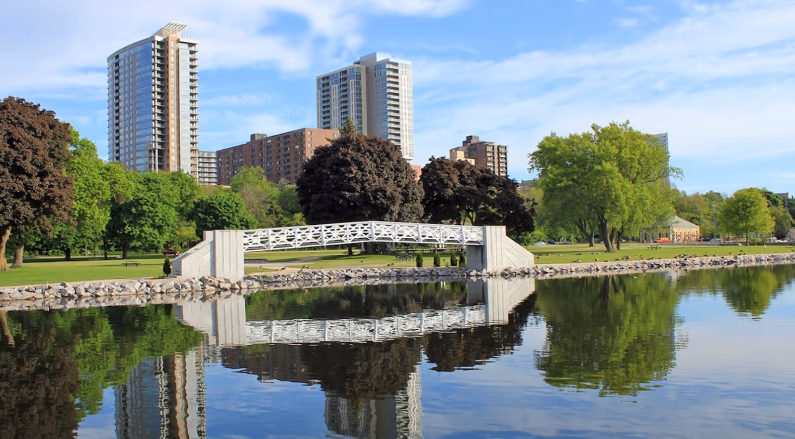 A serene park scene with a white pedestrian bridge over calm water reflecting the bridge, trees, and nearby tall buildings under a blue sky with scattered clouds.