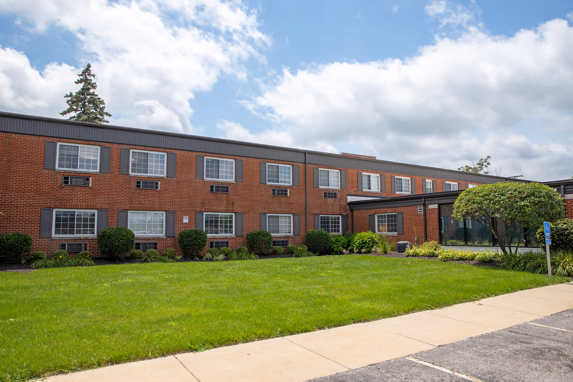 Exterior view of a two-story brick building with multiple windows, surrounded by green grass, shrubs, and a small tree under a partly cloudy sky.