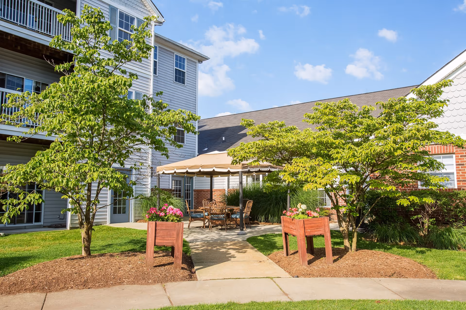Outdoor patio area at Independence Village of White Lake with a paved walkway leading to a shaded seating area with a table and chairs. The patio is surrounded by green trees, flower planters, and well-maintained grass. The building exterior is visible with windows and balconies under a clear blue sky.