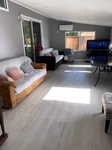 A bright living room with light wood flooring, two sofas with cushions, an armchair, a glass coffee table, a flat-screen TV on a stand, and a window with a view of a wooden fence. The walls are painted gray and there is a sliding glass door on one side.