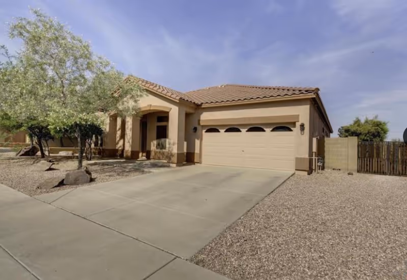 Single-story house with a beige exterior, a tiled roof, a two-car garage, and a driveway. The front yard has desert landscaping with gravel, rocks, and a few trees. The sky is clear with some wispy clouds.