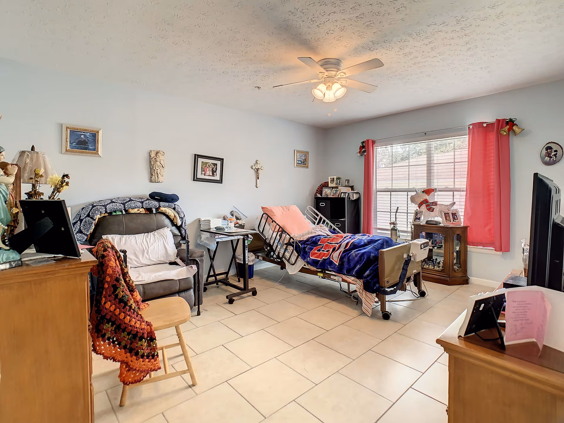 A well-lit bedroom in a senior living facility featuring a hospital-style adjustable bed with a colorful blanket, a recliner chair with a crocheted blanket draped over it, a wooden stool, and various personal items and decorations on the walls and furniture. A large window with red curtains allows natural light to fill the room.