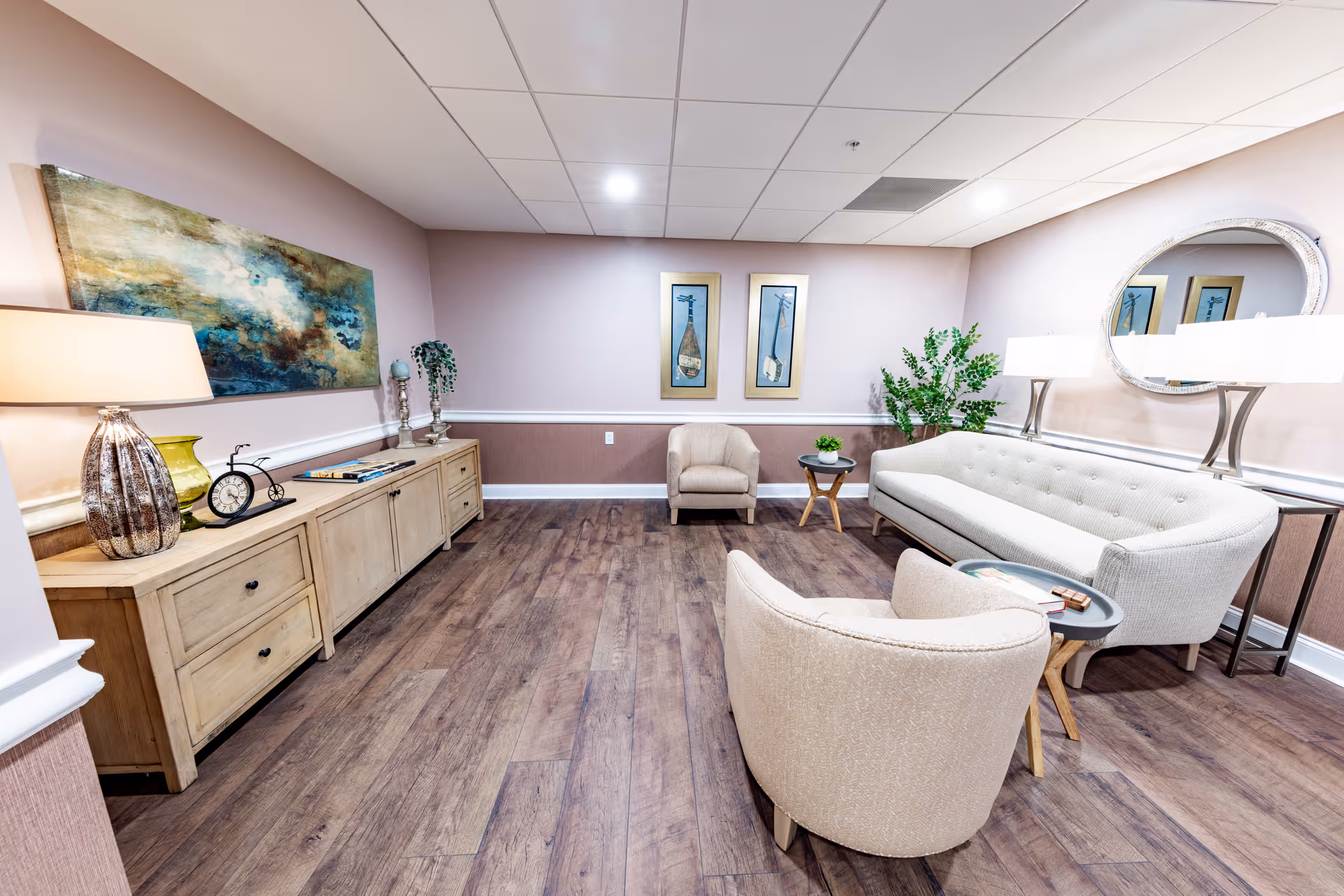 Bright senior living lounge with a sofa and armchairs arranged around side tables, a wooden console with lamps and artwork, and plants on wood flooring.