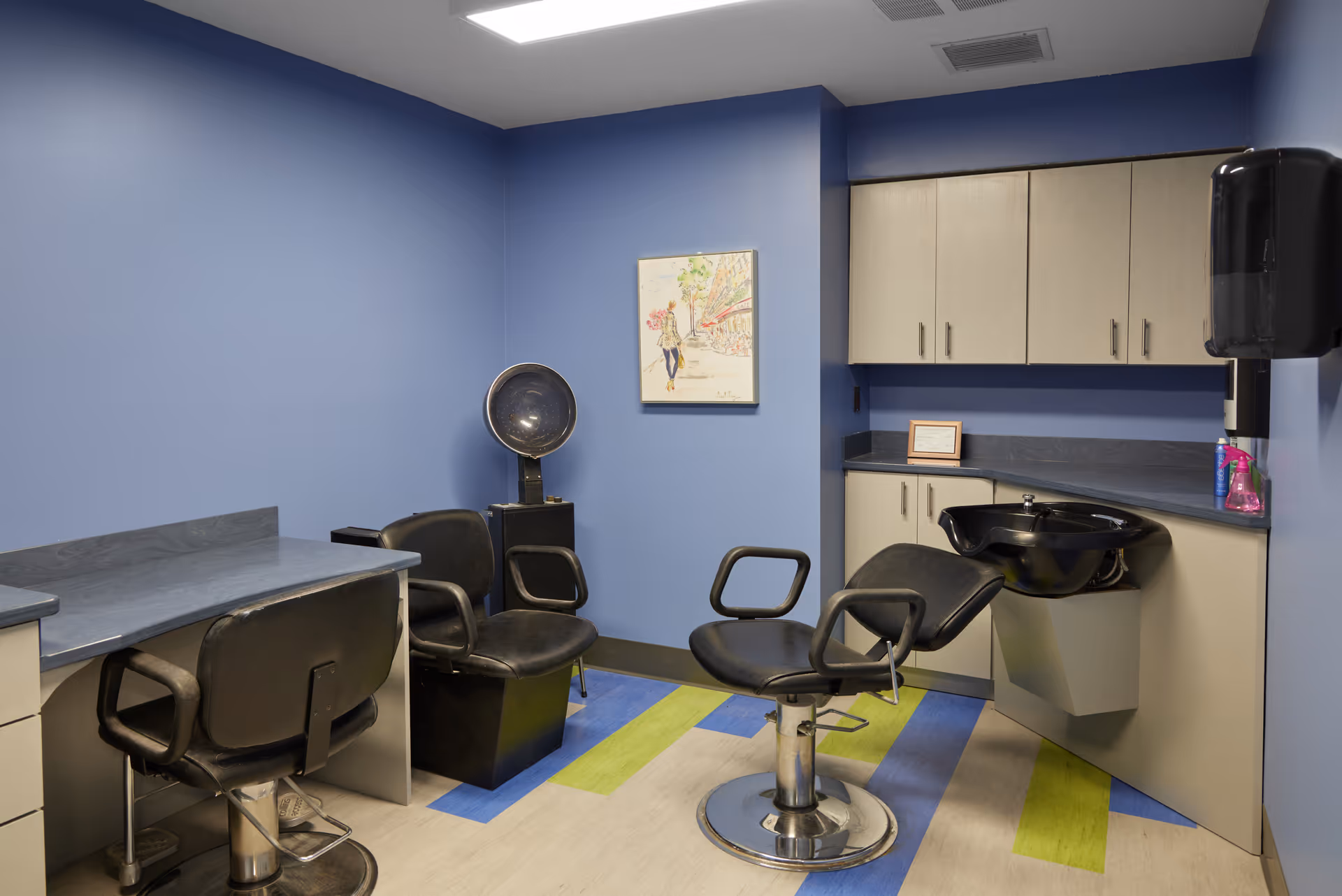 Interior of a salon room with blue walls, featuring two black salon chairs, a hair washing station with a black sink, a desk with a chair, cabinets, and a wall-mounted hair dryer. The floor has a colorful pattern with blue, green, and beige sections, and a framed artwork is hung on the wall.