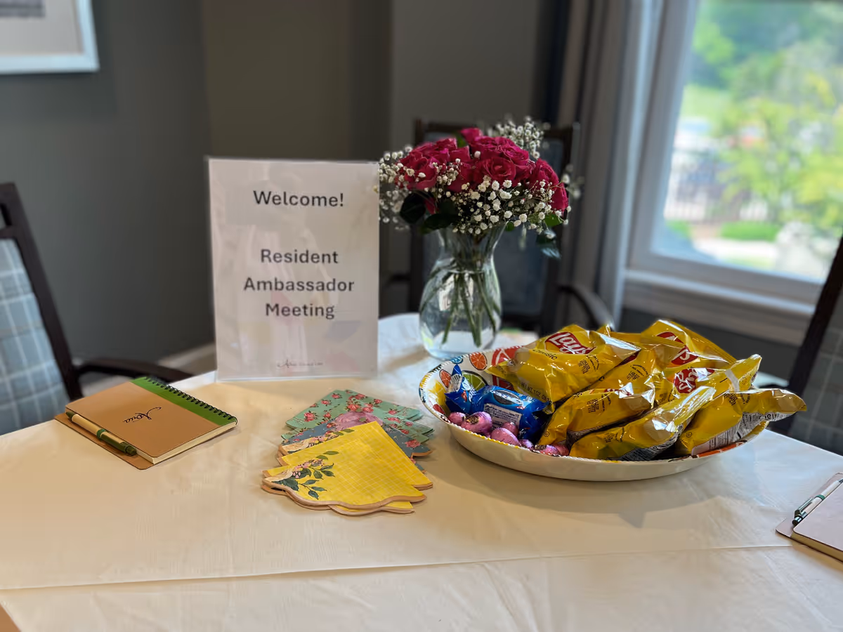 A table set up for a Resident Ambassador Meeting with a sign that says 'Welcome! Resident Ambassador Meeting', a vase of pink roses with white baby's breath, a bowl filled with bags of Lay's chips and other snacks, colorful napkins, a notebook with a pen, and a clipboard. The table is covered with a white tablecloth and is near a window with a view of greenery outside.