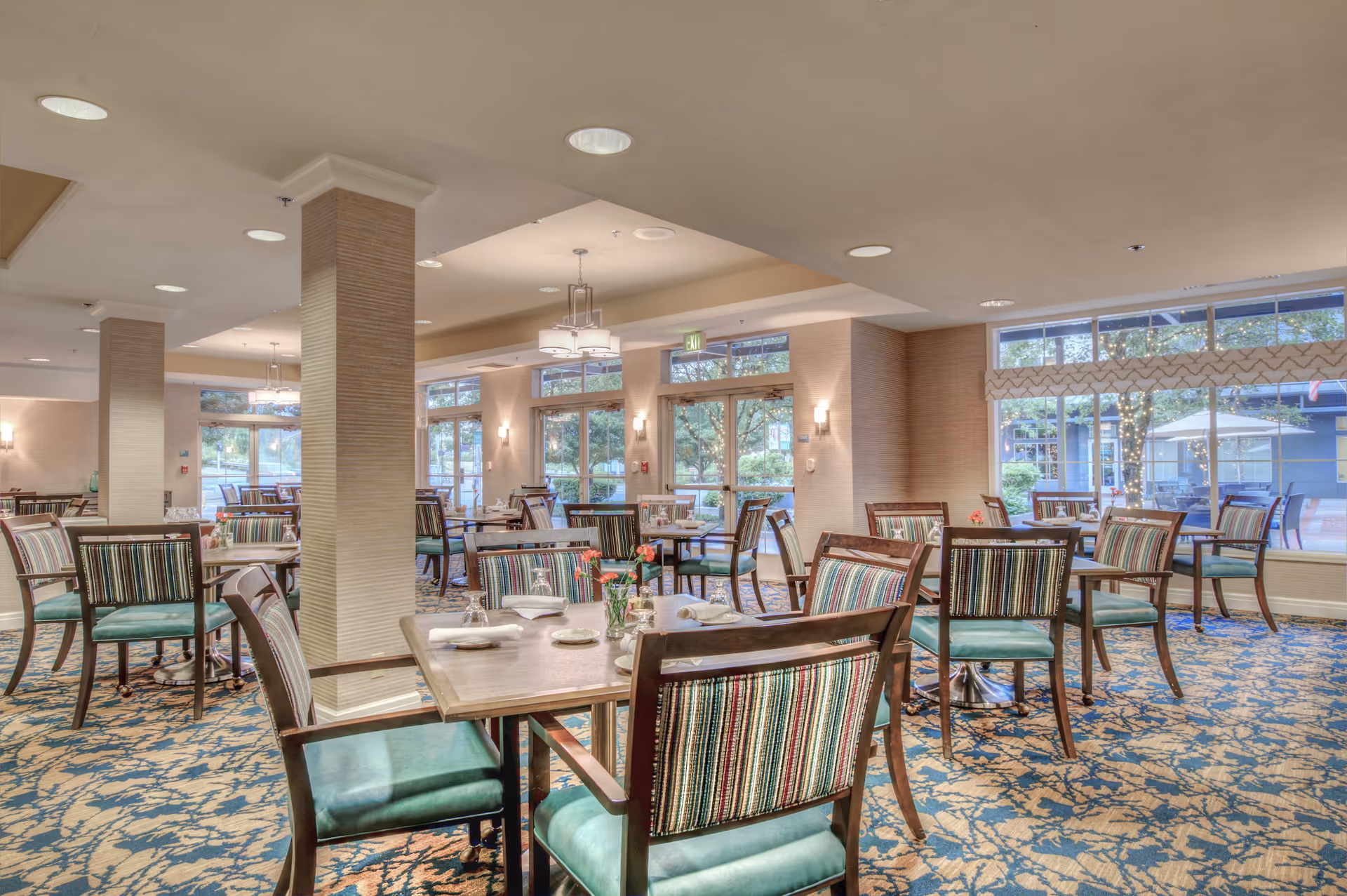 A spacious dining room with multiple wooden tables and chairs featuring striped upholstery and green cushions. The room has large windows letting in natural light, patterned carpet flooring, and modern ceiling lights. There are small flower arrangements on the tables and an outdoor patio area visible through the windows.