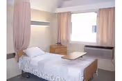 A single hospital-style bed with white bedding and a pillow in a small, simple room. The bed has a wooden headboard and footboard, and there is a wooden bedside table with a small flower vase on it. A window with light pink curtains is on the wall behind the table, and a wall-mounted light fixture is above the bed. There is also a wall-mounted air conditioning unit below the window.