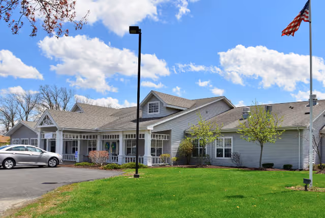 Exterior view of a single-story senior living facility building with gray siding and a gabled roof under a partly cloudy sky. There is a green lawn in front, a flagpole with an American flag on the right, a black street lamp in the center, and a silver car parked on the left side near the entrance.
