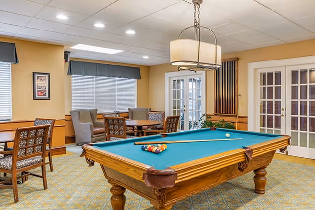A recreational room with a pool table in the center, set up with billiard balls and a cue stick. The room has beige walls, carpeted floor, and several chairs and tables around the perimeter. There are two large windows with blinds and valances, a hanging light fixture above the pool table, and French doors on the right side.