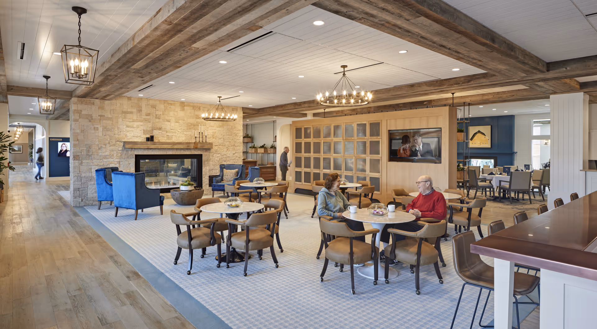 A spacious and well-lit common area in a senior living facility featuring several round tables with chairs, a stone fireplace with blue armchairs, and a large flat-screen TV mounted on a wooden partition. Two elderly people are seated at one of the tables having a conversation. The room has wooden beams on the ceiling, modern chandeliers, and a mix of carpet and hardwood flooring. In the background, there is a dining area with more tables and chairs.