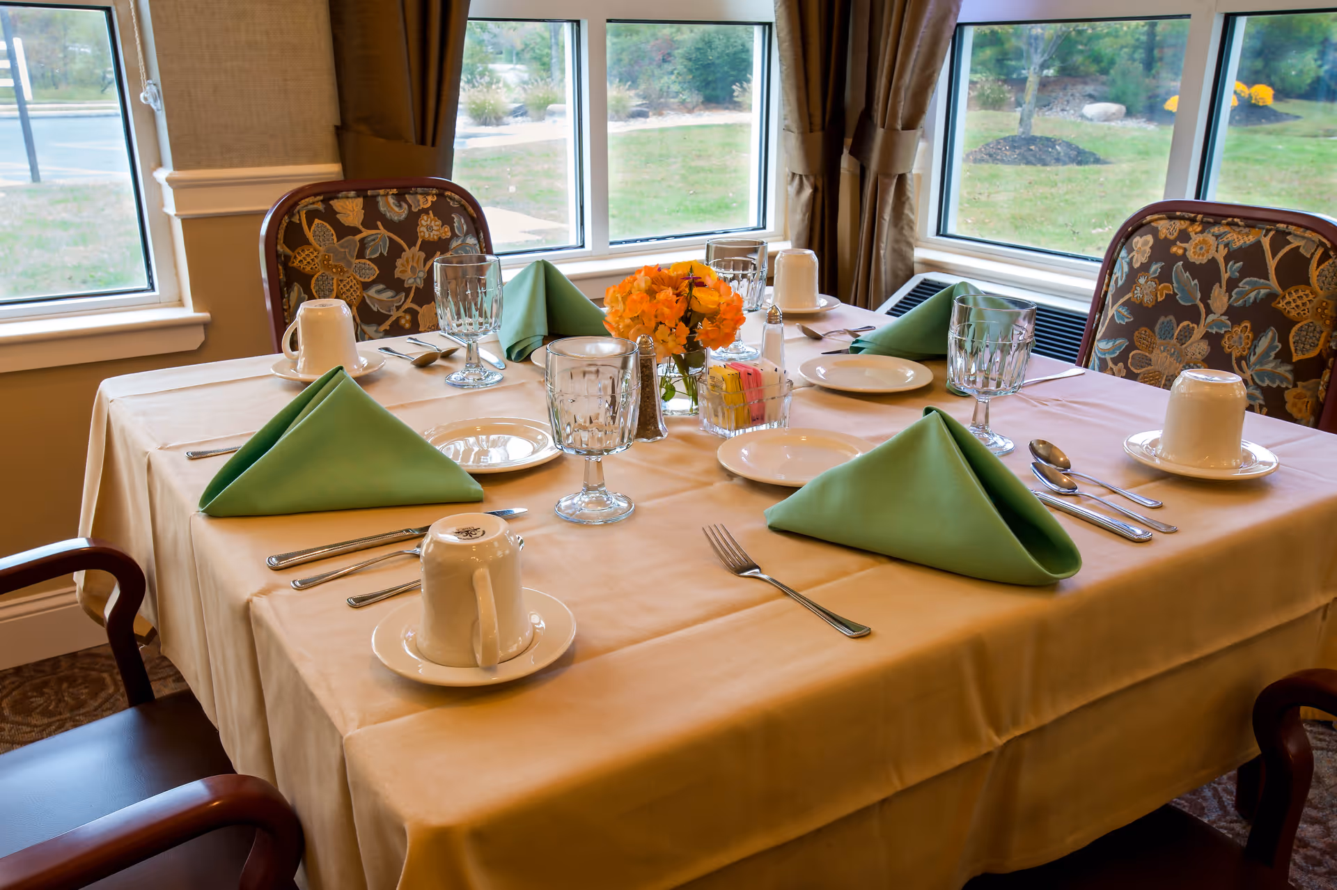 A dining table set for four with beige tablecloth, green folded napkins, white cups on saucers, clear water glasses, silverware, a small vase with orange flowers, and a container with sugar packets. The table is near large windows with a view of greenery outside.