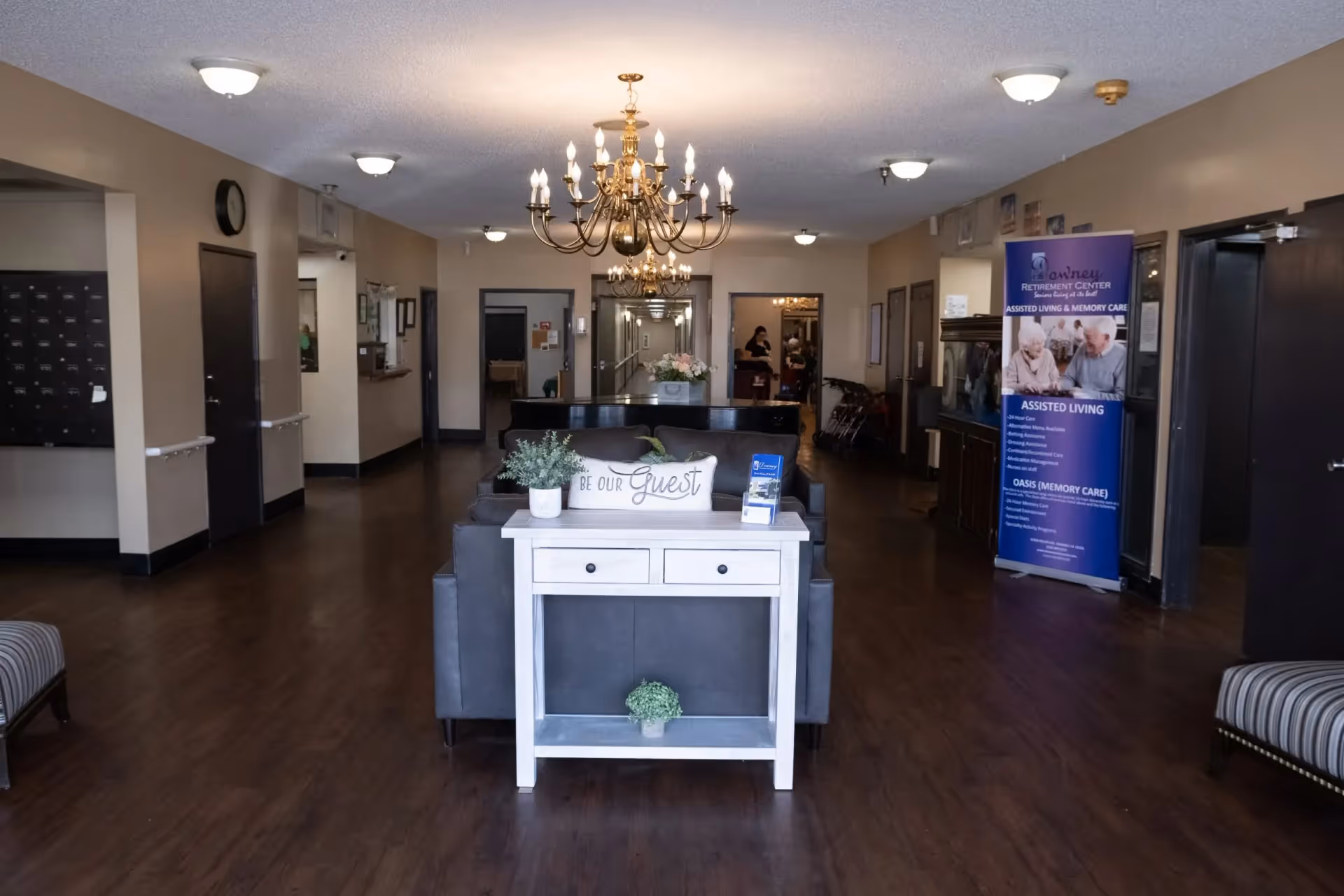 Interior view of a senior living facility lobby with wooden flooring, beige walls, and ceiling lights. A white console table with plants and a 'Be Our Guest' pillow is placed in front of a gray sofa. A chandelier hangs from the ceiling. In the background, there are doorways and a blue informational banner about assisted living and memory care services.