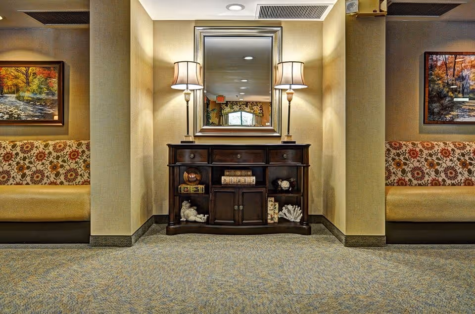 A cozy interior hallway area with a dark wooden console table centered against a beige wall. On the table are two lamps with beige shades and decorative items including books and small sculptures. Above the table is a large framed mirror reflecting the opposite side of the hallway. On either side of the table are built-in seating areas with floral patterned cushions and framed autumn landscape paintings hanging above them. The floor is carpeted in a muted pattern.